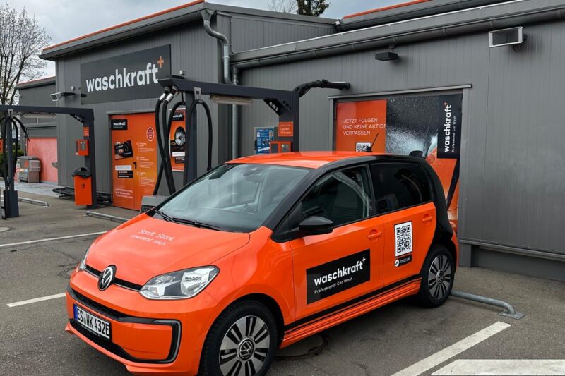 Orangefarbenes Auto mit waschkraft Logo steht vor einer grauen Waschanlage mit derselben Beschriftung.