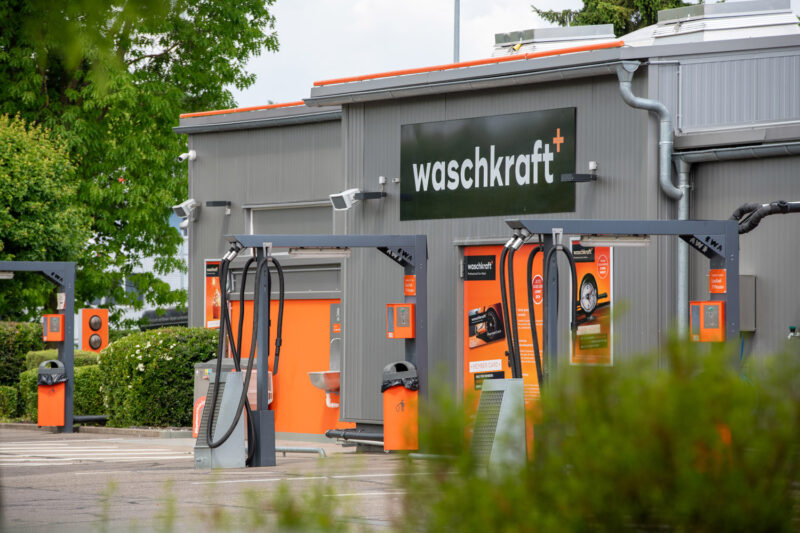 Außenansicht einer Waschanlage von Waschkraft mit mehreren Waschplätzen und einem Gebäude im Hintergrund, umgeben von grünen Büschen und Bäumen.