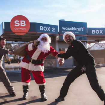 Drei Männer bei einer SB-Autowaschanlage, einer als Weihnachtsmann verkleidet, alle fröhlich posierend.