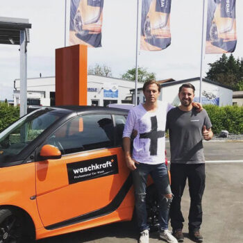 Zwei Männer stehen vor einem orangefarbenen Auto mit "waschkraft" Logo, im Hintergrund wehen Fahnen von waschkraft.