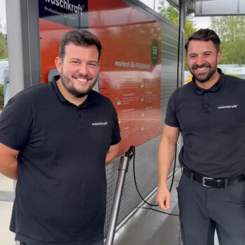 Zwei Männer in Waschkraft-Uniformen lächeln vor einem Waschprogramm-Schild an einer Selbstbedienungs-Autowaschstation.