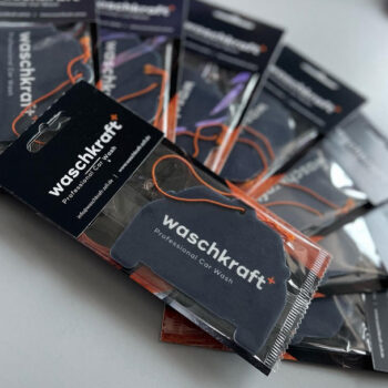 Lufterfrischer von waschkraft in Autoform, verpackt in durchsichtiger Plastikfolie mit orangefarbenen Bändern.
