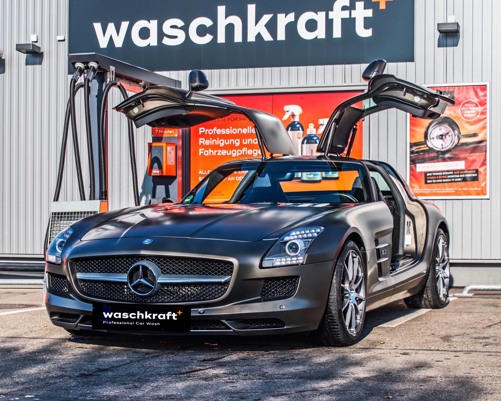 Ein silbergrauer Sportwagen mit geöffneten Flügeltüren steht vor einer professionellen Autowaschanlage namens waschkraft.