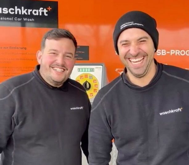 Zwei Männer in schwarzen Pullovern mit waschkraft Logo lächeln vor einem orangefarbenen Hintergrund mit Waschanlagenschalter.