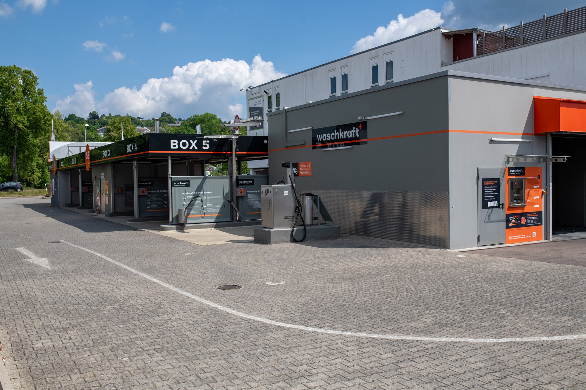 Waschstraße von waschkraft mit Boxen für Autowäsche, graues Gebäude mit orangefarbenen und schwarzen Details, umgeben von Bäumen unter blauem Himmel.