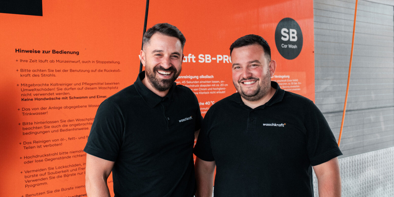 Zwei Männer in schwarzen Waschkraft-Poloshirts stehen lächelnd vor einer orangefarbenen Infotafel in einer SB-Waschanlage.