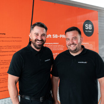 Zwei Männer in schwarzen Waschkraft-Poloshirts stehen lächelnd vor einer orangefarbenen Infotafel in einer SB-Waschanlage.