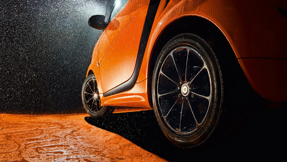 Nahaufnahme eines orangefarbenen Autos während einer Autowäsche, Wassertropfen spritzen auf den lackglänzenden Lack und das Rad.