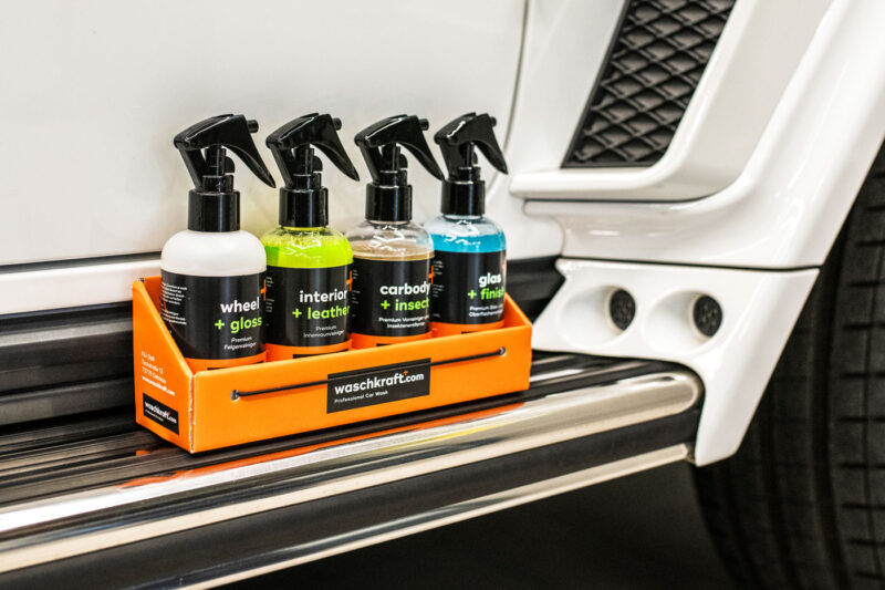 Vier Waschkraft Autopflegeprodukte in Sprühflaschen auf einer orangefarbenen Ablage vor einem weißen Auto.